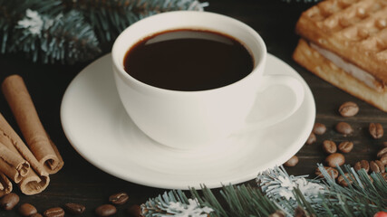 White cup of hot coffee on a brown wooden table. Green spruce branches, homemade waffles with souffle, cinnamon and coffee grains. Festive cozy mood. New Year and Christmas concept. Depth of field