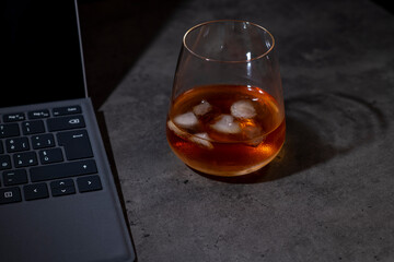 whiskey glass with ice next to laptop on dark gray cement background