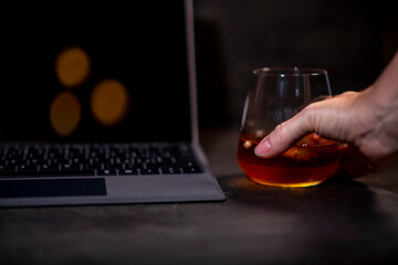 whiskey glass with ice in hand next to laptop on dark gray cement background