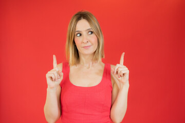 Portrait of a happy young woman dressed in red dress pointing fingers up at copy space isolated over red background