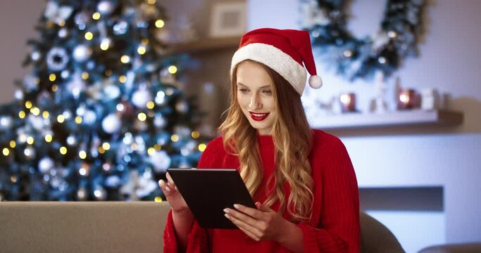 Portrait of joyful young beautiful Caucasian woman in santa hat in decorated modern room with xmas tree tapping and scrolling on tablet and choosing xmas gifts on Christmas Eve. Festive mood concept