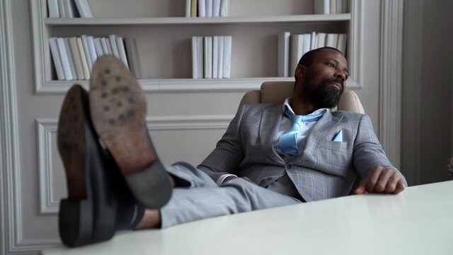 millionaire is boring in his working office, sitting in chair with legs on table, handsome black rich man is resting