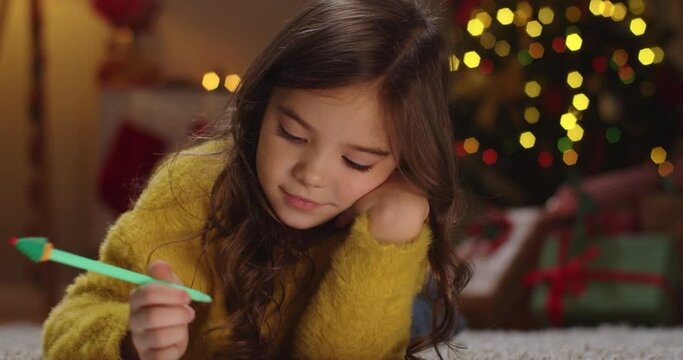 Close up portrait of Caucasian joyful cute little girl laying in decorated room on floor drawing on paper with smile on face. Writing letter to Santa Claus. Winter holidays. Christmas concept