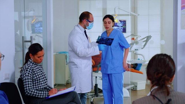 Dentist showing teeth x-ray reviewing it with nusre. Doctor and assistant working in modern crowded stomatological clinic, patients sitting on chairs in reception filling in dental forms and waiting