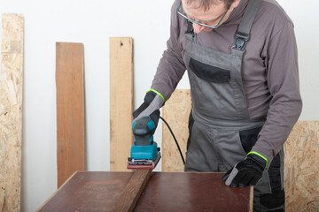 Carpenter working with  electric sheet finishing sander  on wood lath on desk, on background oriented strand board