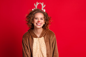 cute funny young woman in a Christmas reindeer costume smiles and rejoices on colorful red background.