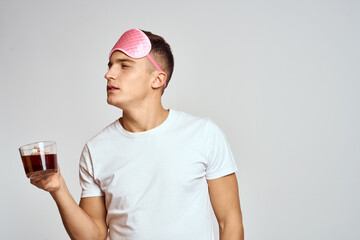 handsome man with pink sleep mask and with a cup of tea in hand pulls himself up on a light background