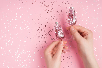 Close up top above overhead first person view photo of female hands holding two toy tree baubles champagne glasses over pastel pink color background with glitter