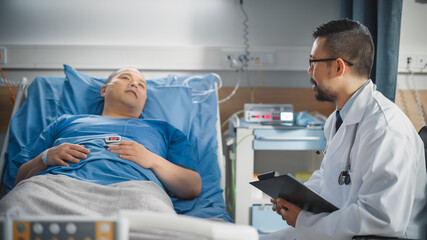 Fototapeta premium Hospital Ward: Friendly Chinese Doctor Sits Near and Talks to Handsome Male Patient Resting in Bed, Explains Test Results. Physician Talks to Man Recovering after Successful Surgery