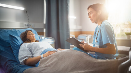 Obraz premium Hospital Ward: Friendly Hispanic Head Nurse Fills Medical History Form, Talks to Female Patient Recovering in Bed. Professional Nurse Helps Happy Woman Get Better after Surgery. Shot with Sun Flare.