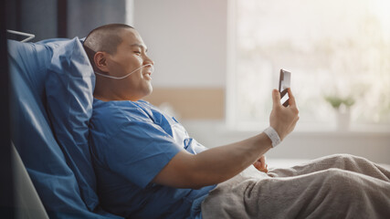 Medical Hospital Ward: Portrait of Handsome Chinese Man with Nasal Cannula Resting in Bed, Uses Smartphone to Video Chat with His Family, Friends, Shooting Blog. Surgery Recovery Room. Side View.