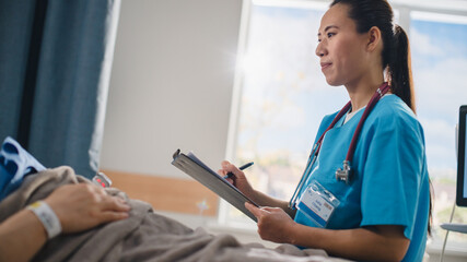 Hospital Ward: Portrait of Friendly Chinese Head Nurse Talks to Anonymous Patient Recovering in Bed and Fills Medical History Form. Doctor Helps Person Get Better. Dutch Angle Shot.