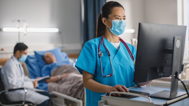 Hospital Ward: Professional Experienced Chinese Head Nurse / Doctor Wearing Face Mask Uses Medical Touch Screen Computer, Checking Patient's Medical Data. In the Background Patient Recovering on Bed.