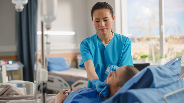 Hospital Ward: Friendly Chinese Head Nurse Adjusts Nasal Cannula to a Sick Male Patient Resting in Bed According Treatment Plan. Nurse Helps Man Getting well after Serious Surgery.