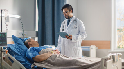 Hospital Ward: Friendly Chinese Doctor Talks to Handsome Male Patient Resting in Bed, Uses Tablet Computer, Explains Test Results. Physician Talks to Man Recovering after Successful Surgery