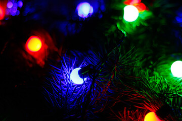 New Years blurred background with light lamps on fir spruce branch. Coloured round light. Flashlight in the dark. Texture fir. Festive mood. Holiday dark night background. Selective focus