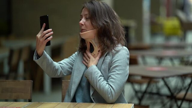 woman with orthopedic collar on neck is communicating by video call on mobile phone in cafe, curing osteochondrosis