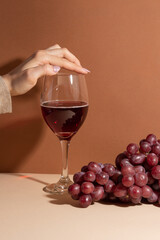 Tender woman finger touch the wineglass with red wine with branch of grapes background. Beige surface.