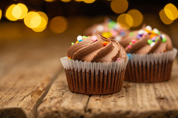 Close-up view of tasty chocolate cupcakes with sprinkling on rustic wooden table on black background with yellow lights. Sweet dessert. Bakery concept. Elegant food. Sweets for coffee or tea.