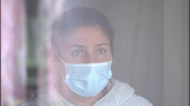 Young caucasian woman in a coronavirus mask touching with her hand to the glass of the window. Sad woman in quarantine. Pensive young woman in mask to protect herself from covid-19 while looking out