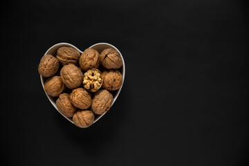 Whole walnuts and one cracked broken heart in a white heart-shaped bowl on a black background. Top view. Place for text.
