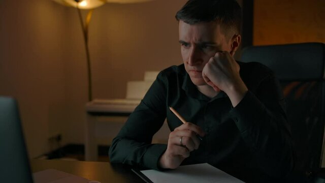 Caucasian male thinks about solving hard mathematic equation in online studying. Handsome student writes notes in text pad and look at laptop screen in night mood interior.