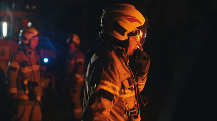 Fototapeta na wymiar Close up of firefighter using radio to speak with his collegues. Fire engine with emergency lights and two more firemen in the background. High quality photo