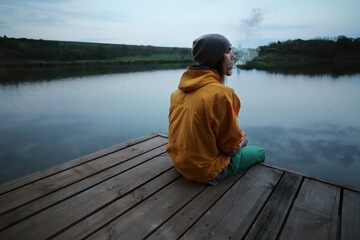 Young sports male addict smokes marijuana near the river at sunset. - obrazy, fototapety, plakaty