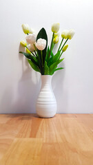 Fake tulip and green leaves in white vase on wooden table with wall background. Ornamental flower for decoration home, house or place indoor. Fresh petal or flora.