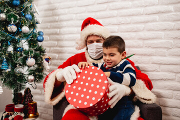 Excited cheerful boy in a striped T-shirt sitting in Santa's lap with a face mask and opening a red xmas gift next to the Christmas tree. Surprise for a child during the COVID-19 coronavirus pandemic