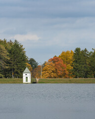 a vertically oriented medium close shot of a small building on a lake with multicolored fall leaves on the trees in the background