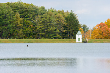 wide angle landscape shot of a small white building sitting on the far side of a lake with the New England fall foliage in the background 