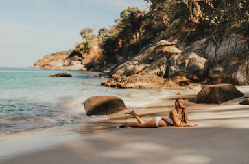 Fototapeta na wymiar young blond European woman in white bikini swimsuit on beach lies on sand beach sea. background of large stone rocks and palm trees