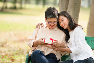 Young beautiful Asian woman smiling and gives warm hug with a present to her mom in the park as a background, Happy new year gift, concept mothers day, mother-daughter love, Special day for mom.