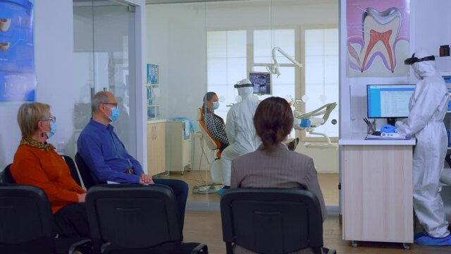 Patients with protection masks wating doctor in dental clinic reception while stomatologist working in background wearing ppe suit. Concept of new normal dentist visit in coronavirus outbreak.
