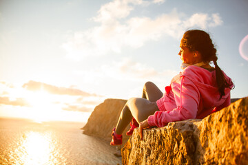 Back view of woman sitting on a mountain top admires the rising sun over the sea or ocen and enjoys...