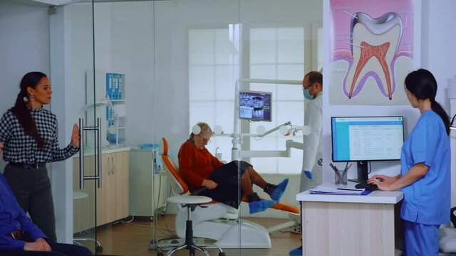 Dentist inviting senior man in consultation dental room while nurse giving to patient form to fill indicating to sit on chair in waiting area. Slow motion shot crowded professional orthodontist office