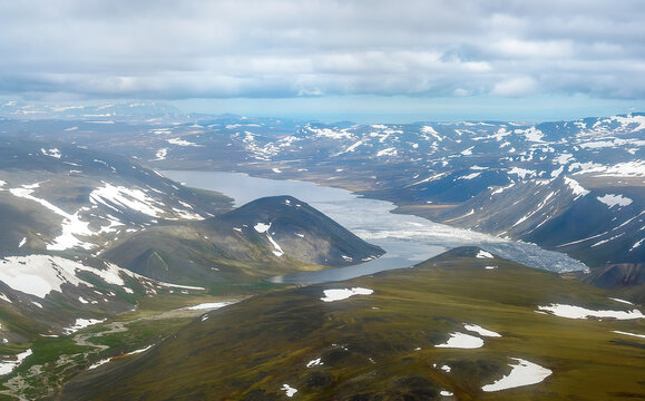 Aerial view of the lake among the mountains. Summer arctic landscape. Snow in the tundra and on the mountain slopes. Nature of Chukotka and Siberia. Lake Koolen, Inan Range, Chukotka, Far East Russia.