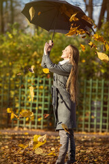 Attractive happy woman in a gray coat holds an umbrella in her hands. Leaf fall in the autumn park. Beautiful young woman is walking among the falling leaves. Yellow foliage. Fall season. Autumn mood.