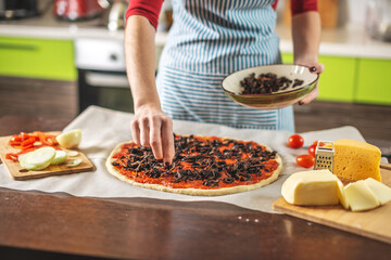 Female chef in an apron is putting mushrooms on a pizza raw. Cooking delicious pizza at home in the kitchen.