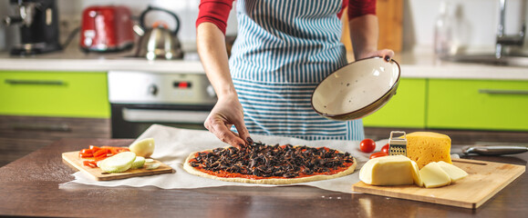 Female chef in an apron is putting mushrooms on a pizza raw. Cooking delicious pizza at home in the kitchen.
