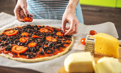 Female chef in an apron is putting the tomatoes on a raw pizza. Cooking delicious pizza at home in the kitchen