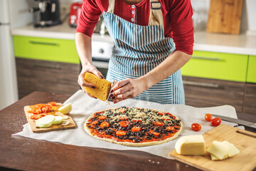 Female chef in an apron is rubbing cheese on a raw pizza. Cooking delicious pizza at home in the kitchen.