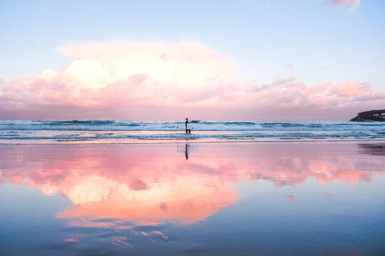 Side View Of Man With Surfboard Walking On Shore At Beach Against Dramatic Sky During Sunset