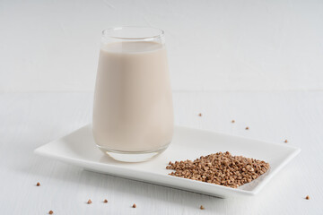 Gluten-free and lactose-free vegetarian buckwheat milk full of vitamins, minerals and antioxidants served in tall drinking glass with raw grains on plate on white wooden background. Horizontal