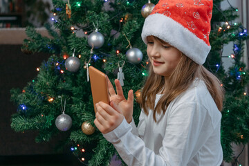 Online New Year and Christmas congratulations. Cute smiling girl uses mobile phone for online video chat, call to parents, family or friends