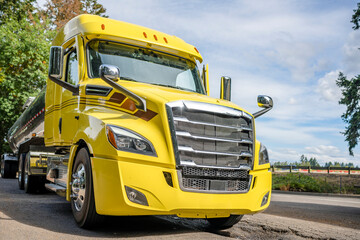 Big rig yellow semi truck with shiny tank semi trailer standing on rest area parking lot with old trees