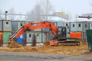 A large orange excavator on wide iron tracks stands on a construction site on the sand against the backdrop of temporary housing for builders