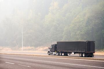 Black stylish big rig semi truck with covered dry van semi trailer transporting cargo driving on the road in the smog from a forest fire
