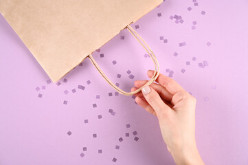 Close up female hold in hand empty craft paper gift bag for purchases after shopping on pastel violet background. Packaging template mockup. Delivery concept advertising mock